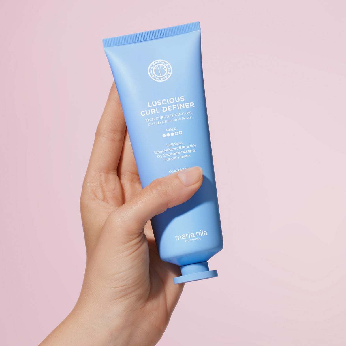 Eine Hand hält eine blaue Tube des maria nila Coils & Curls Luscious Curl Definer vor einem hellrosa Hintergrund.