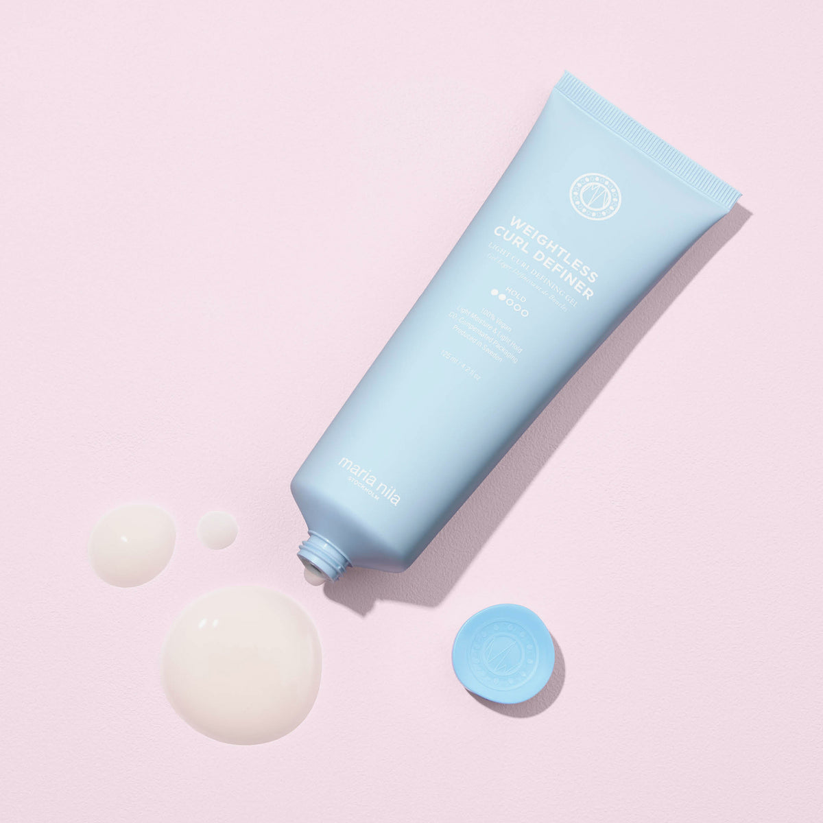 Geöffnete blaue Tube maria nila Coils & Curls Weightless Curl Definer mit Cremeklecksen liegt auf einer hellrosa Fläche.