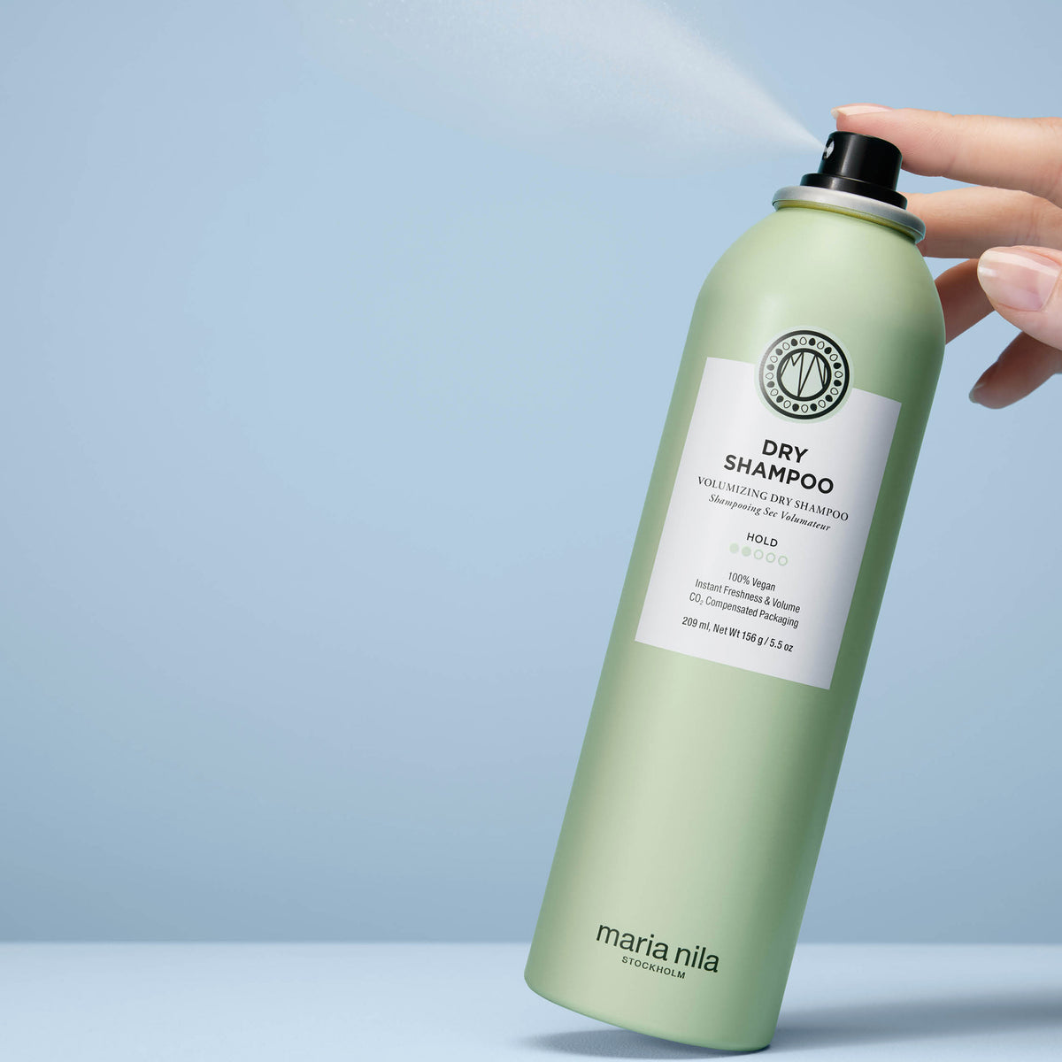 Eine Hand sprüht maria nila Dry Shampoo   aus der grünen Sprühflasche vor einem hellblauen Hintergrund.