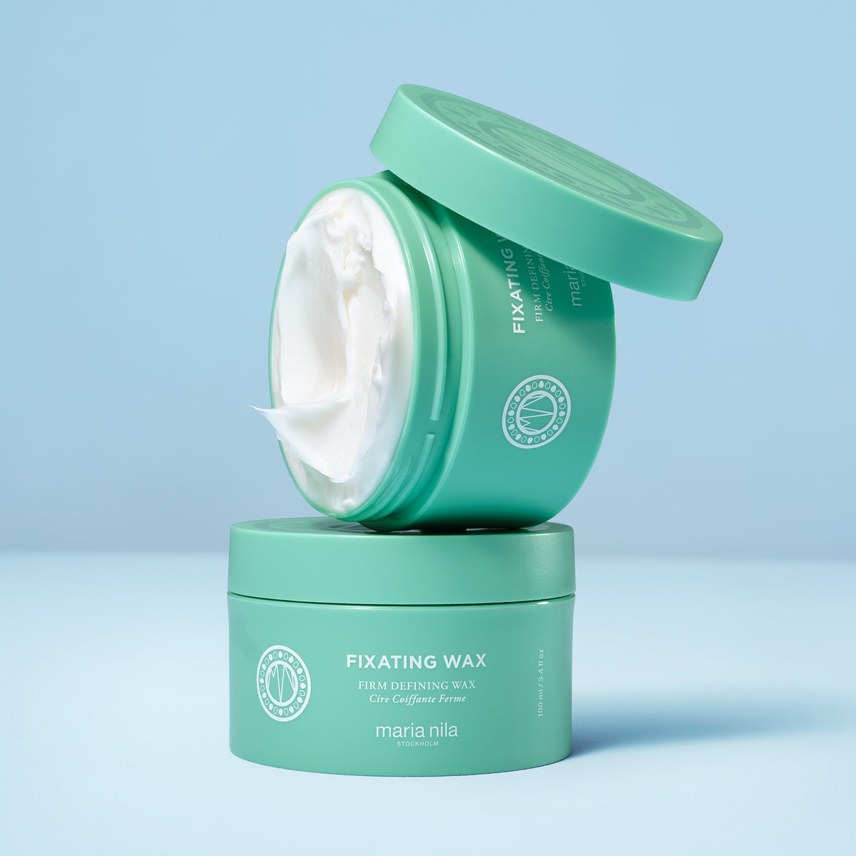 Zwei grüne Tiegel mit maria nila Fixating Wax sind vor einem blauen Hintergrund abgebildet, wobei ein Tiegel geöffnet ist und die weiße Creme im Inneren zum Vorschein kommt.