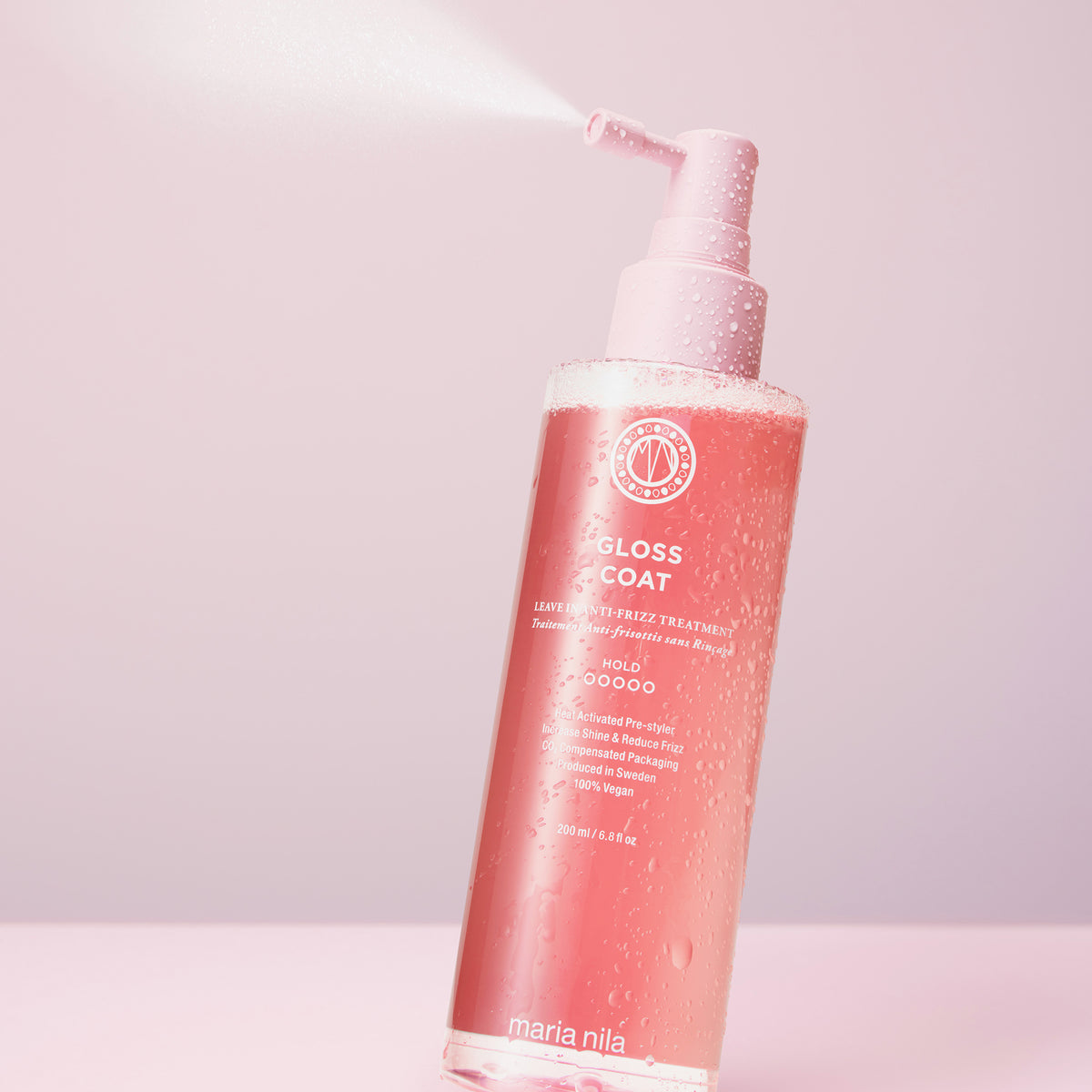 Eine Flasche maria nila Gloss Coat mit Wassertropfen und Sprühnebel auf einem rosa Hintergrund.
