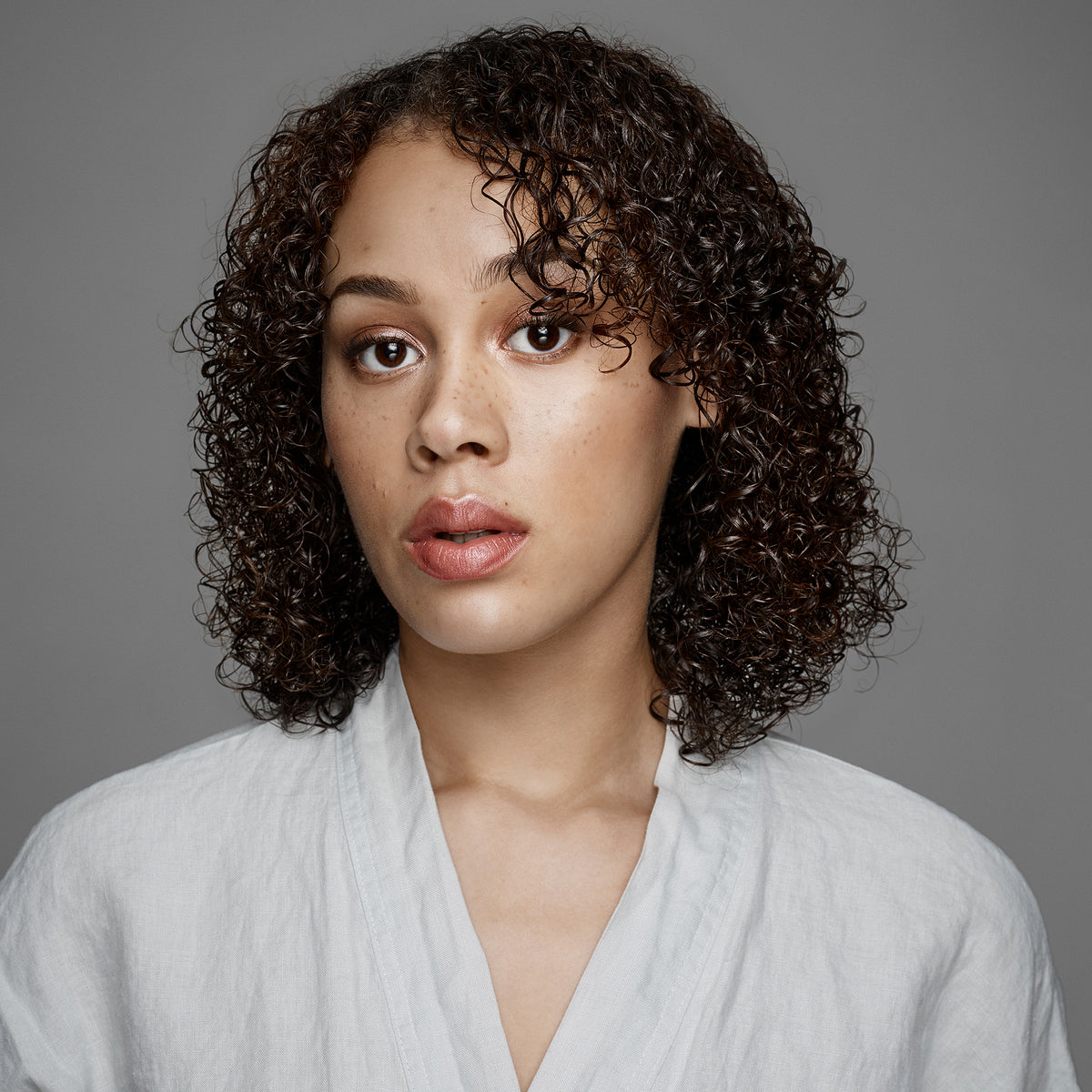 Eine junge Frau mit lockigem braunem Haar und neutralem Gesichtsausdruck zeigt gesunde Locken, die mit maria nila Head & Hair Heal Conditioner gestylt wurden. 