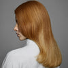 Eine Frau mit glattem, langem, rötlich-blondem Haar vor einem grauen Hintergrund zeigt den gesunden Glanz, der mit maria nila Luminous Colour Conditioner erzielt wurde.