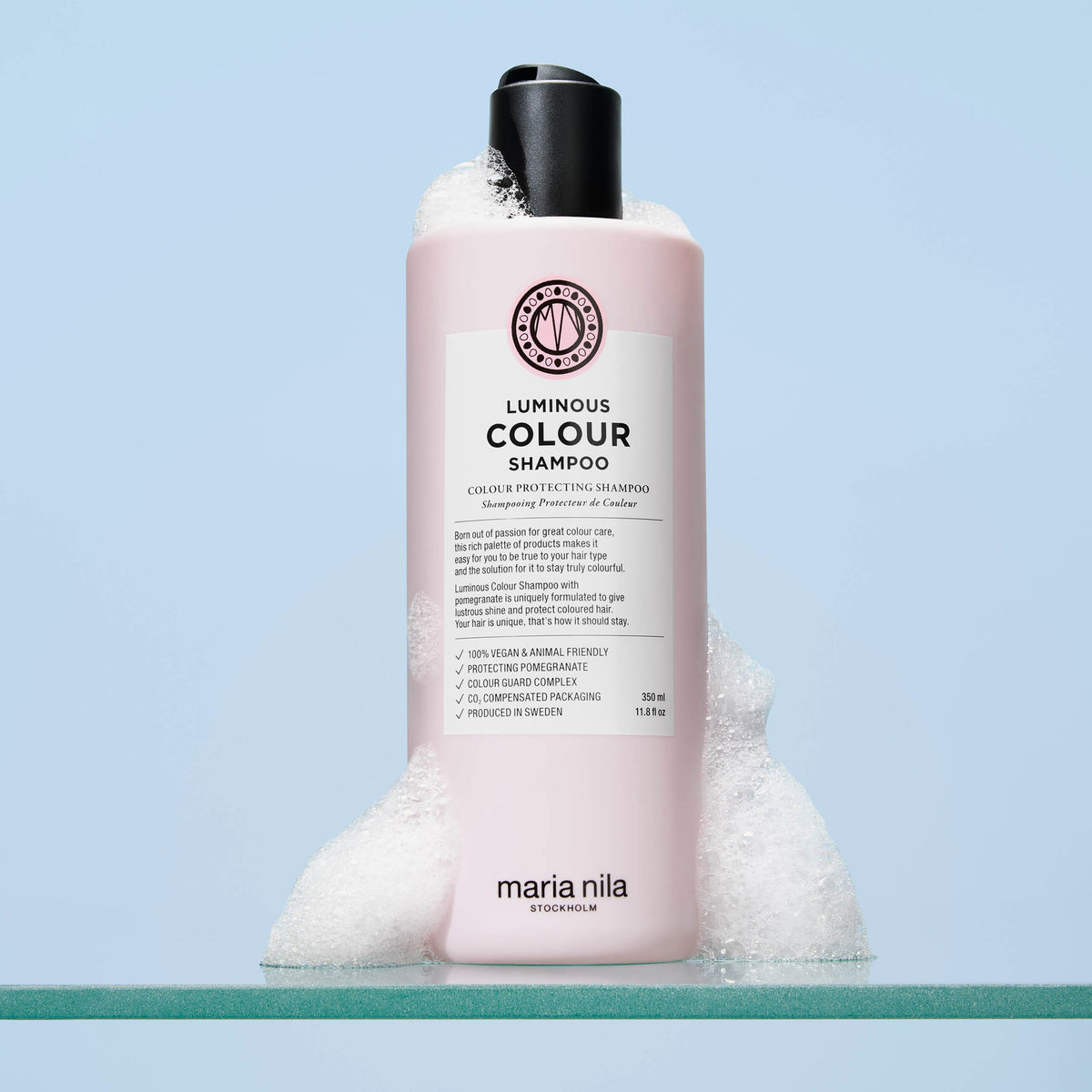 Eine rosafarbene Flasche von maria nila Luminous Colour Shampoo mit Schaum steht vor einem blauen Hintergrund.