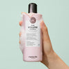 Eine Hand hält eine schaumbedeckte rosa Flasche maria nila Pure Volume Shampoo vor einem tealfarbenen Hintergrund.