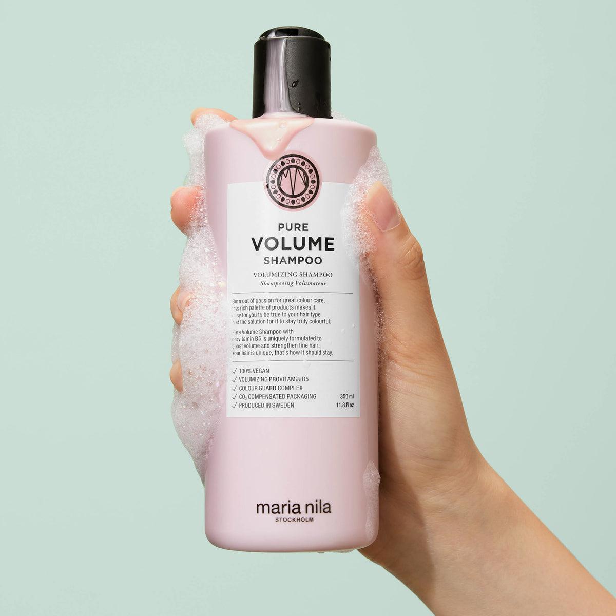 Eine Hand hält eine schaumbedeckte rosa Flasche maria nila Pure Volume Shampoo vor einem tealfarbenen Hintergrund.