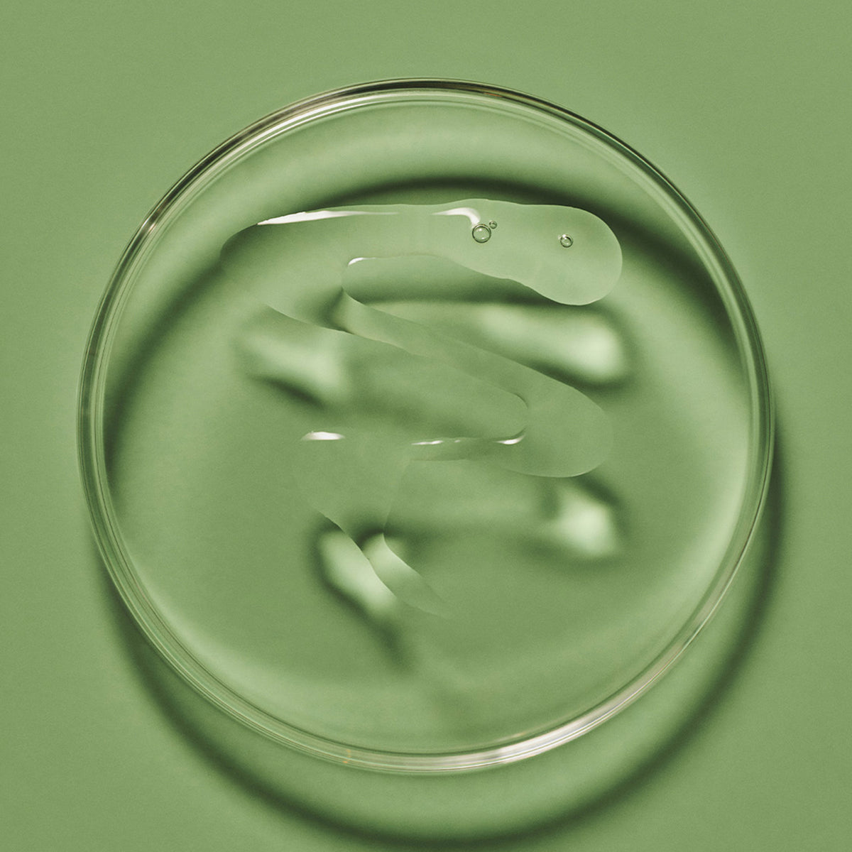 Eine klare Flüssigkeit auf einer Petrischale auf grüner Oberfläche, die das Purifying Cleanse Exfoliating Serum von Maria Nila präsentiert.
