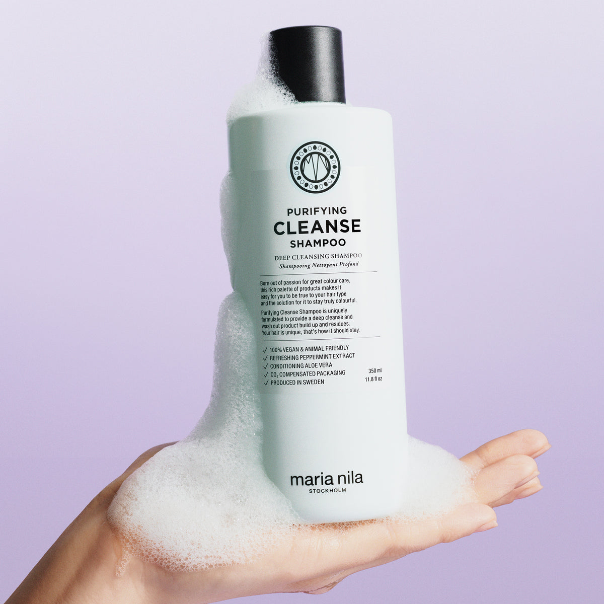 Eine Hand hält eine schaumige Flasche des maria nila Purifying Cleanse Shampoo vor einem hellvioletten Hintergrund.