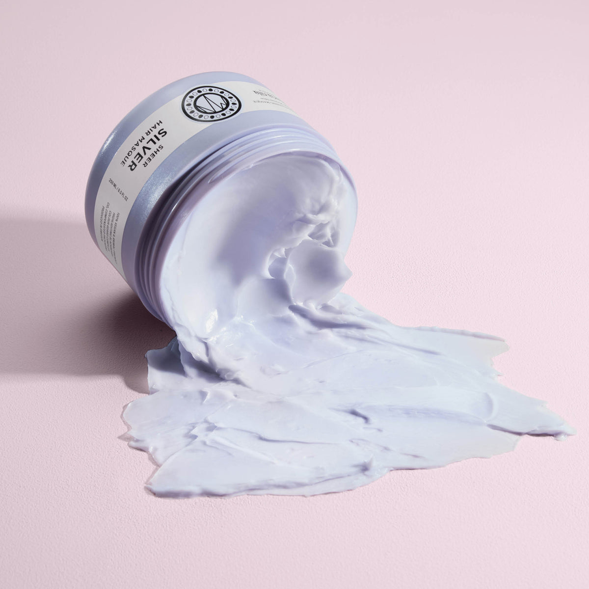 Ein geöffneter Tiegel der lavendelfarbenen maria nila Sheer Silver Masque ergießt sich über eine pastellrosa Oberfläche.