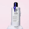 Eine Flasche maria nila Sheer Silver Shampoo mit lila Shampoo und Schaum steht vor einem hellrosa Hintergrund.