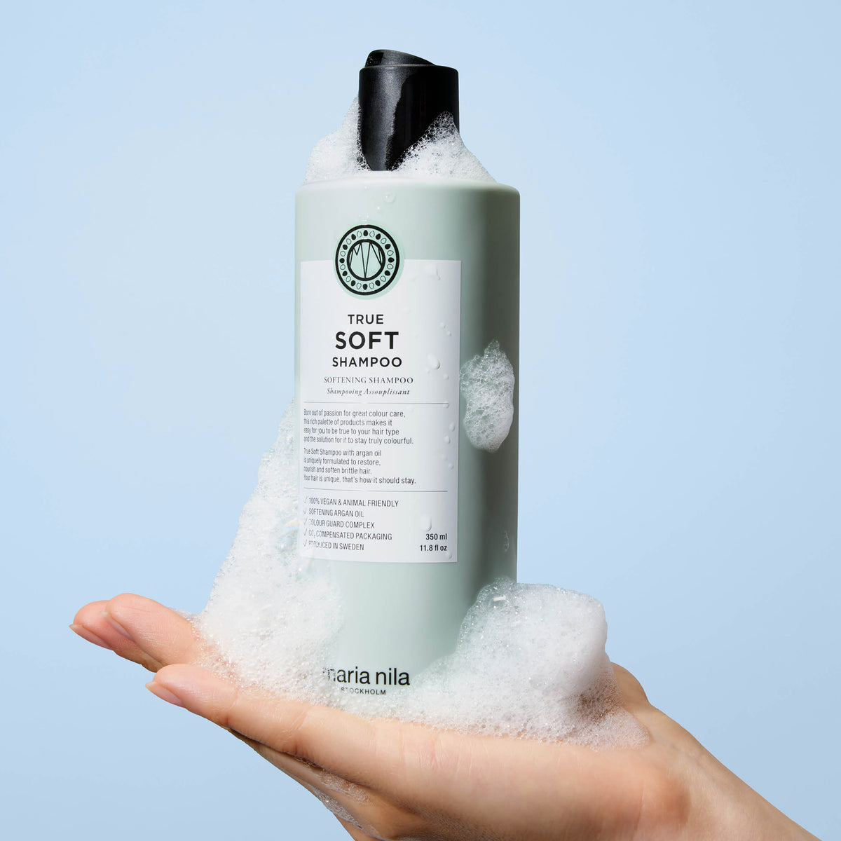 Eine Hand hält eine Flasche maria nila True Soft Shampoo mit weißem Schaum vor einem hellblauen Hintergrund.