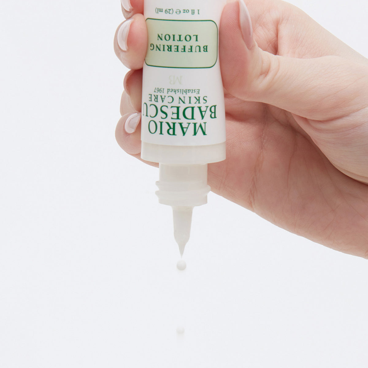 Eine Hand drückt eine Tube Mario Badescu Buffering Lotion zusammen, wobei ein Tropfen aus der Düse austritt.