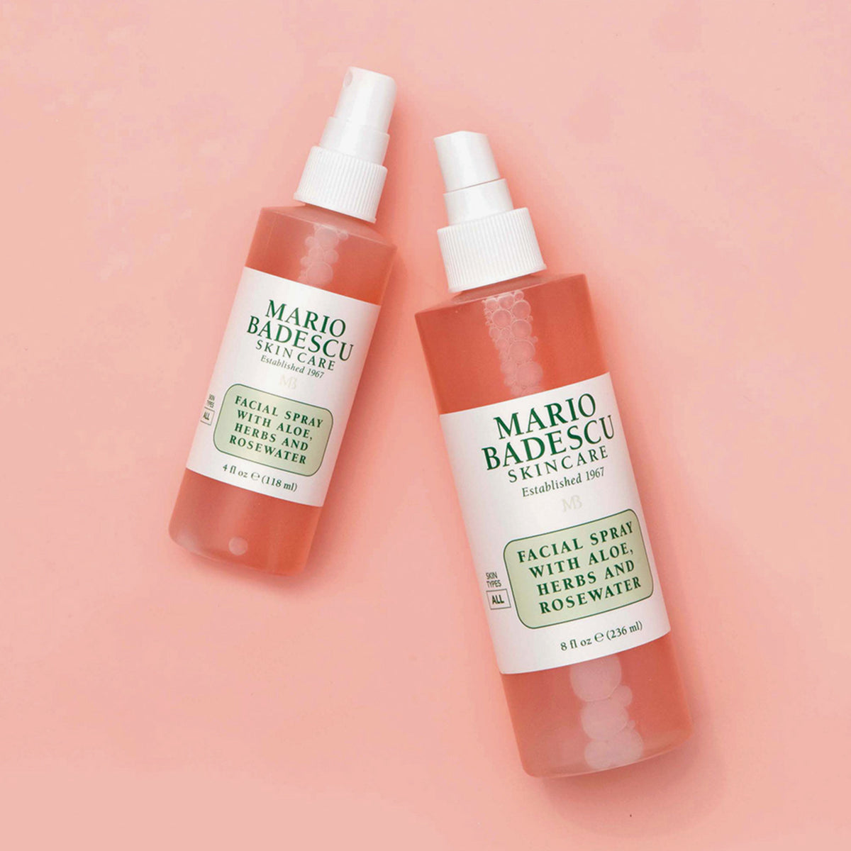 Zwei Flaschen Mario Badescu Gesichtsspray mit Aloe, Kräutern und Rosenwasser sind vor einem rosa Hintergrund abgebildet.