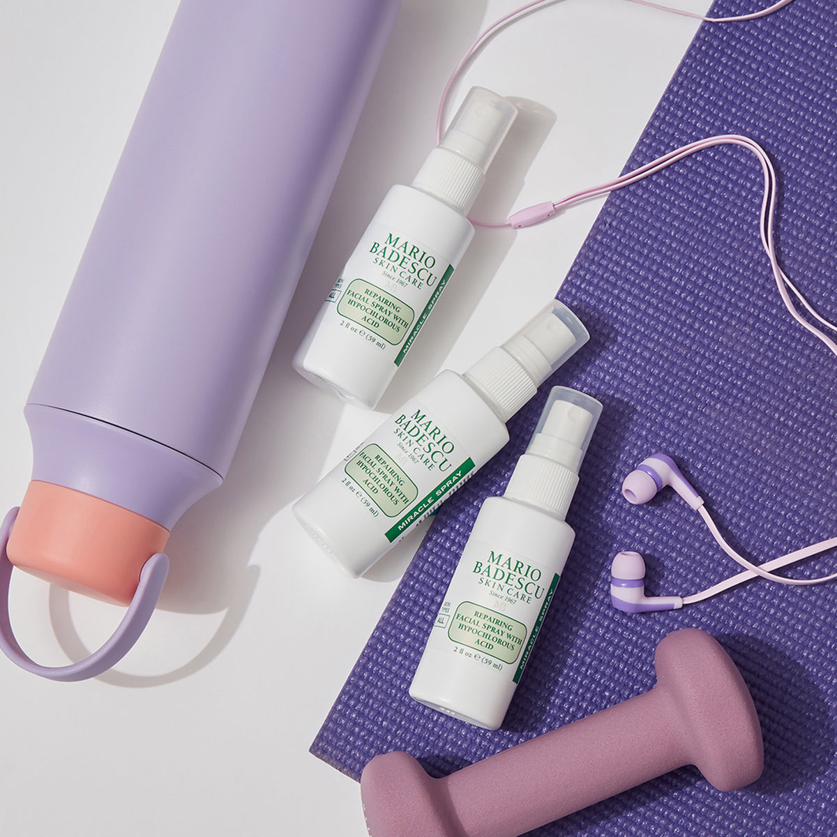 Eine lilafarbene Wasserflasche, eine Trainingsmatte, Kopfhörer, eine Hantel und drei Flaschen Mario Badescu Repairing Facial Spray mit hypochloriger Säure sind nebeneinander angeordnet.