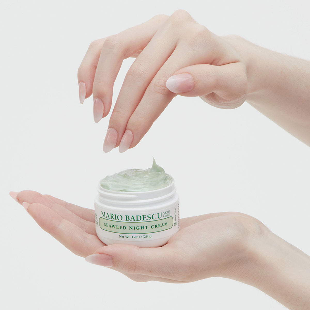 Eine Hand hält einen Tiegel der Mario Badescu Seaweed Night Cream, während die andere Hand in die Creme eintaucht.