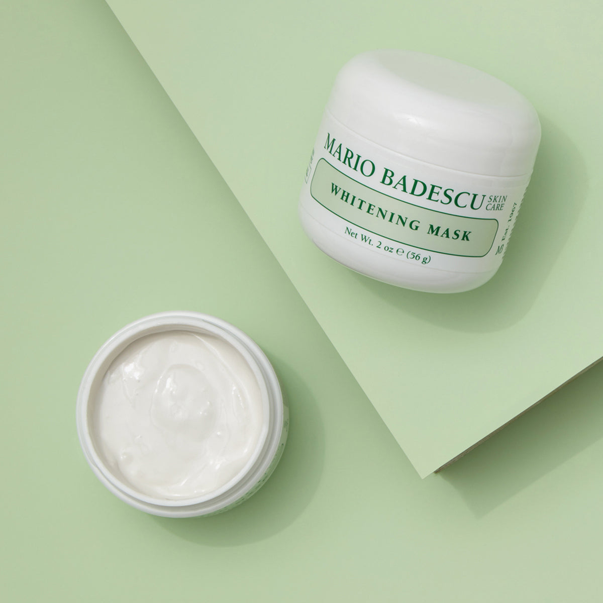Zwei Tiegel der Mario Badescu Whitening Mask stehen auf einem hellgrünen Hintergrund, von denen einer geöffnet ist, um die Creme darin zu zeigen.