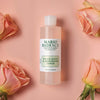 Eine Flasche Mario Badescu Witch Hazel & Rosewater Toner ist zwischen rosa Rosen auf einem pfirsichfarbenen Hintergrund zu sehen.