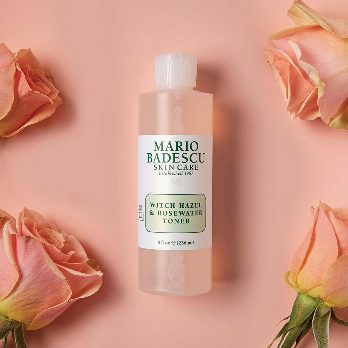Eine Flasche Mario Badescu Witch Hazel & Rosewater Toner ist zwischen rosa Rosen auf einem pfirsichfarbenen Hintergrund zu sehen.