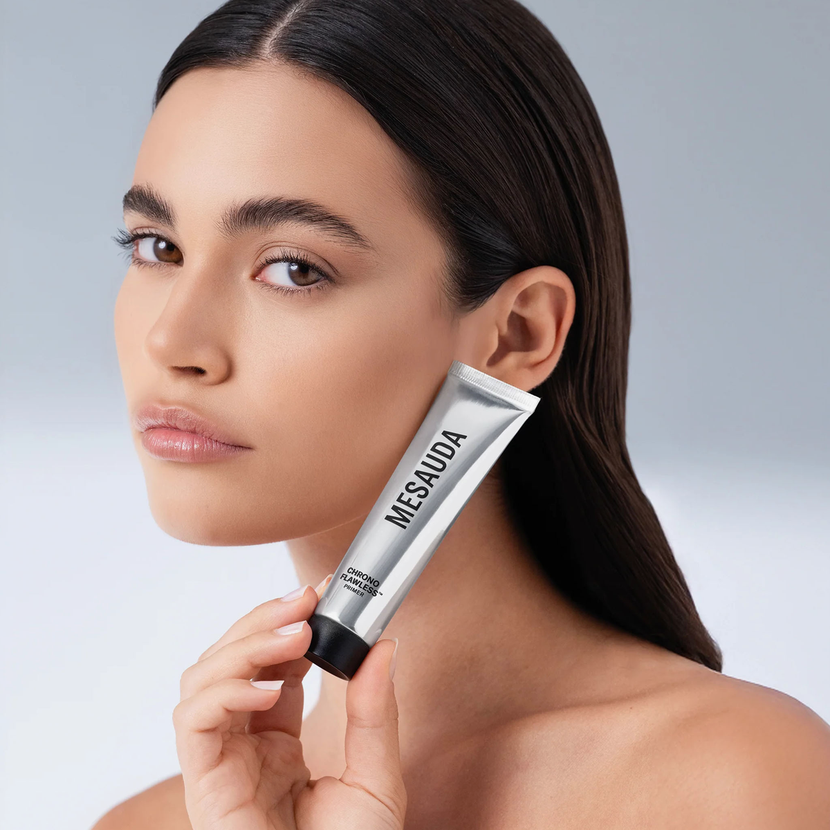 Eine Frau mit glattem braunem Haar hält eine Tube Mesauda Beauty Chrono Flawless Primer in der Nähe ihres Gesichts vor einem grauen Hintergrund.