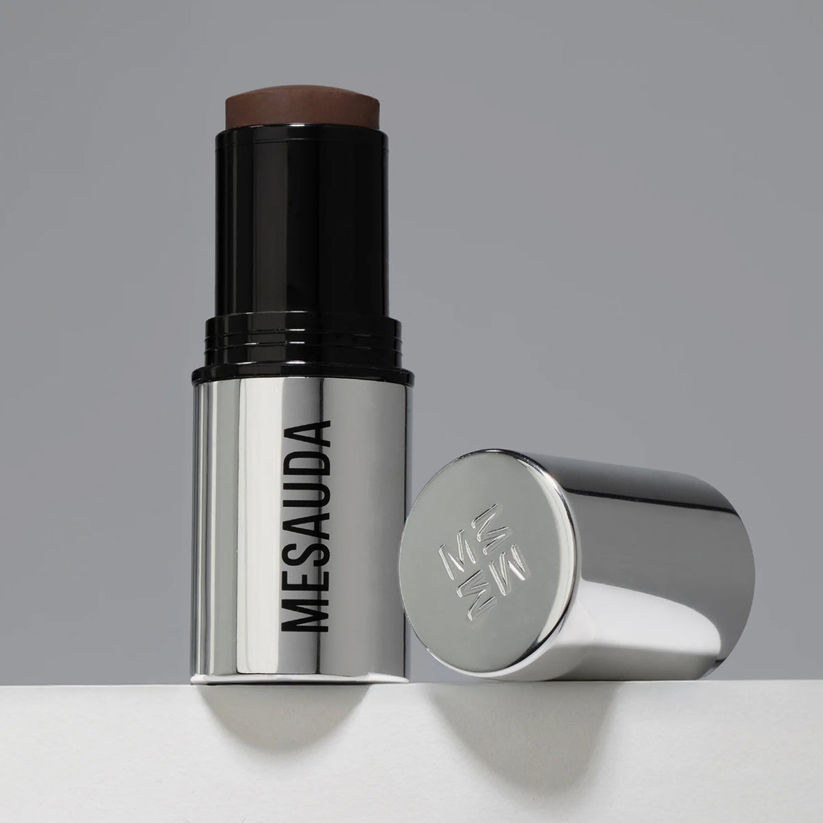 Der silberne Make-up-Stift The Mood Contrast 104 Deep von Mesauda neben silberner Kappe auf grauem Hintergrund.