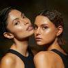 Zwei Frauen mit strahlender Haut posieren eng aneinander in schwarzen Oberteilen, gestylt, und präsentieren Lust For Shine von Mesauda Beauty.