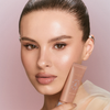Eine Frau mit strahlender Haut hält eine Tube der NABLA Cosmetics Angel Aura Bright Lotion vor einem rosa Hintergrund.