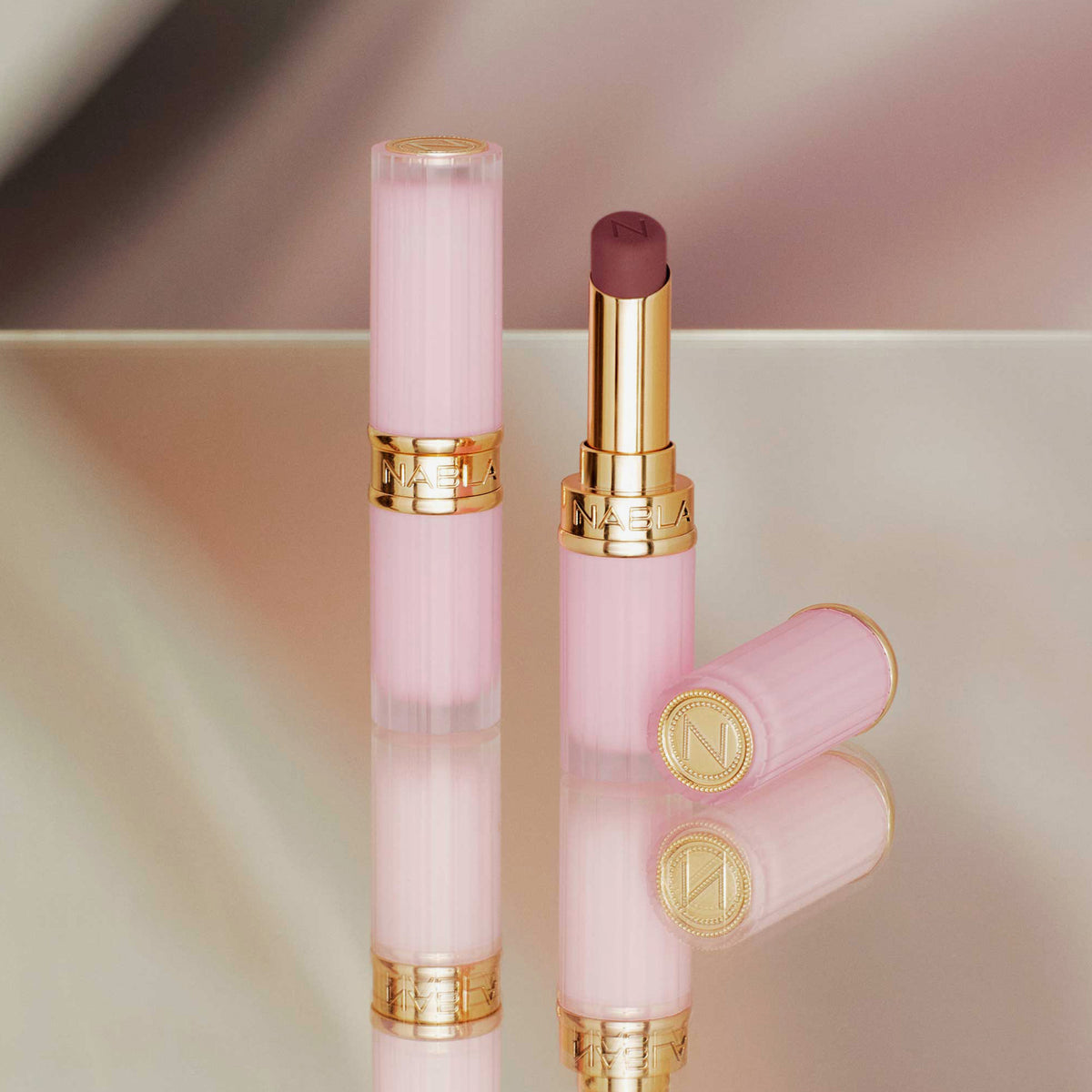 Zwei Beyond Blurry-Lippenstifte in Pink und Gold, einer offen und einer geschlossen, präsentiert auf einer reflektierenden Oberfläche.