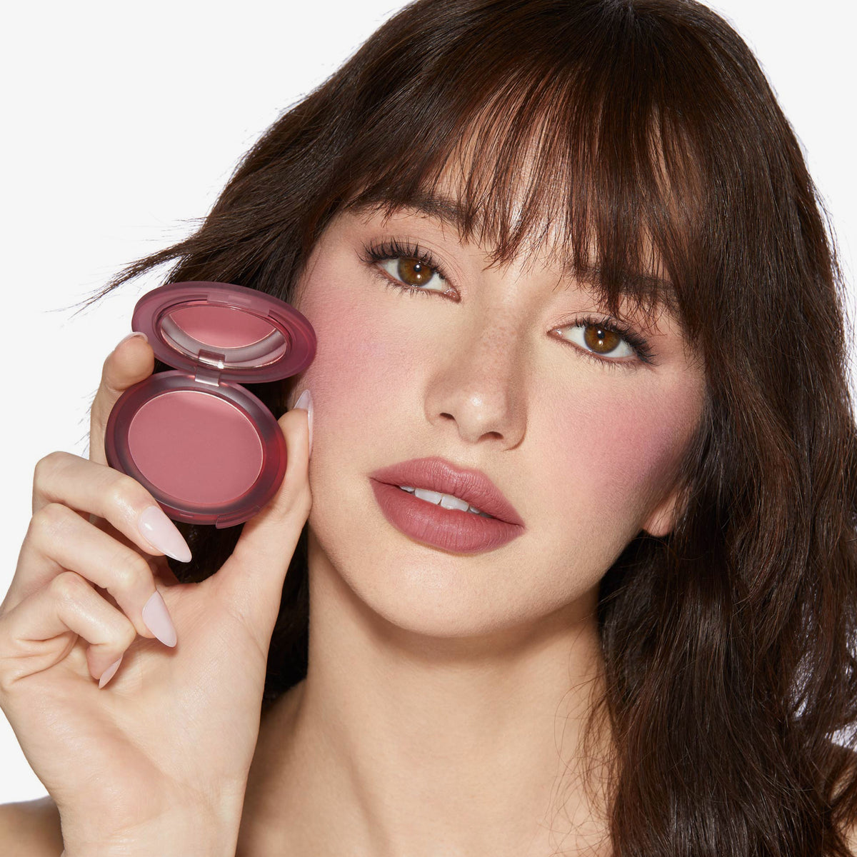 Eine Frau hält ein geöffnetes Close-Up Blurring Powder Blush, trägt den Blush auf den Wangenknochen und natürliches Make-up und blickt in die Kamera.