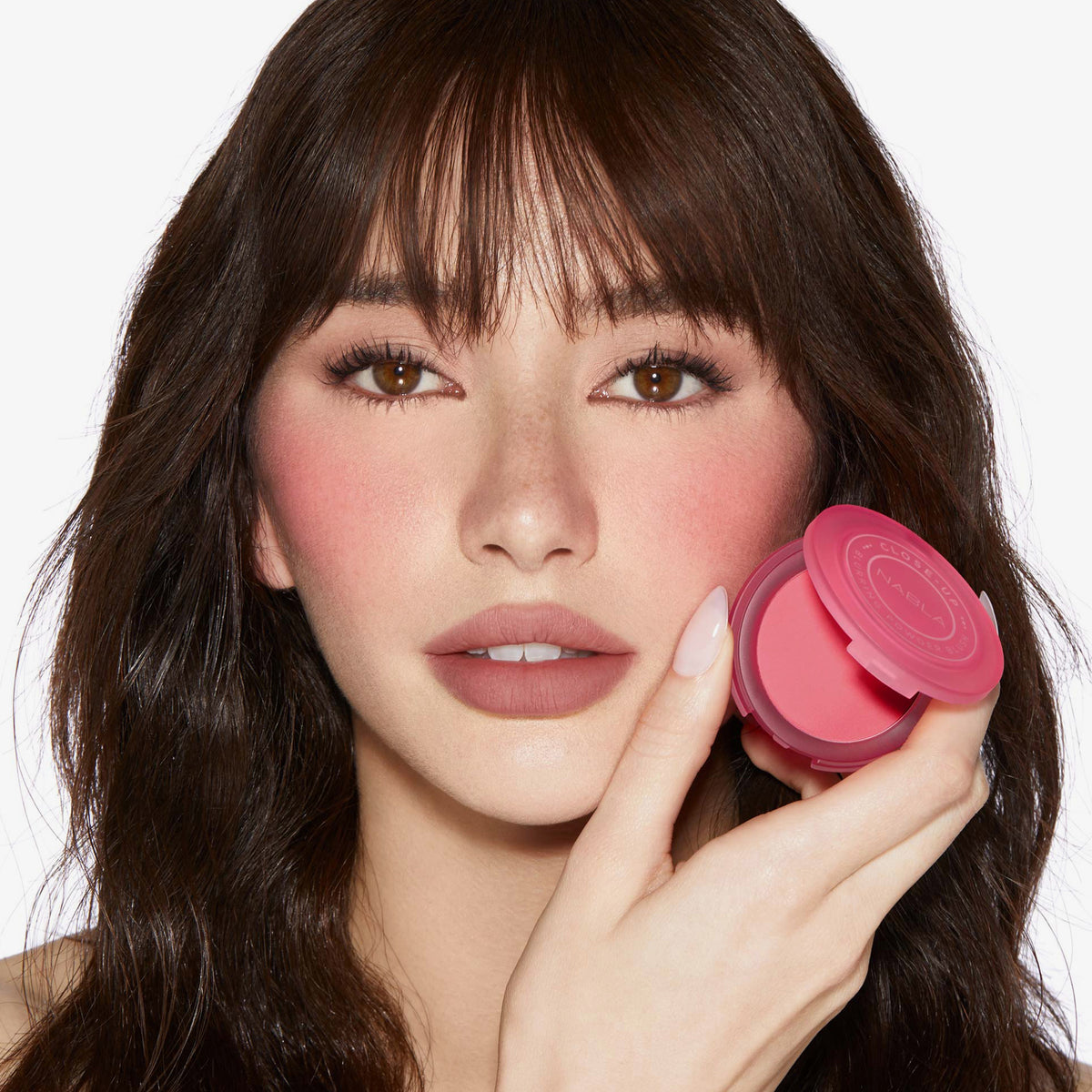Eine Frau mit welligem braunem Haar trägt Close-Up Blurring Powder Blush auf ihren Wangennund ergänzt damit ihren rosigen Make-up-Look.