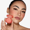 Eine Frau mit kräftigen Augenbrauen hält vor einem weißen Hintergrund das Close-Up Blurring Powder Blush Compact an ihr Gesicht und trägt es auch auf ihren Wangen.