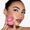 Eine Frau mit kräftigen Augenbrauen hält eine geöffnete Kompaktpackung Close-Up Blurring Powder Blush an ihre Wange.