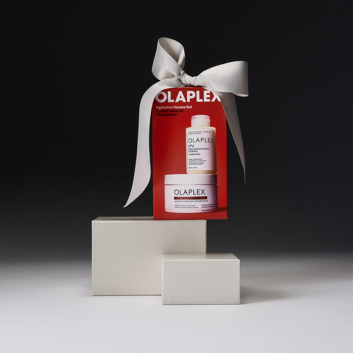 Das OLAPLEX Hydration Heroes Holiday Kit 2026-Geschenkset enthält eine Shampooflasche und einen Tiegel mit Haarmaske, die mit einem cremefarbenen Band an zwei Blöcken befestigt sind.
