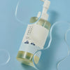 Flasche des 1025 Dokdo Cleansing Oil von Round Lab auf blauem Untergrund, umgeben von geschwungenen, transparenten Ölklecksen.