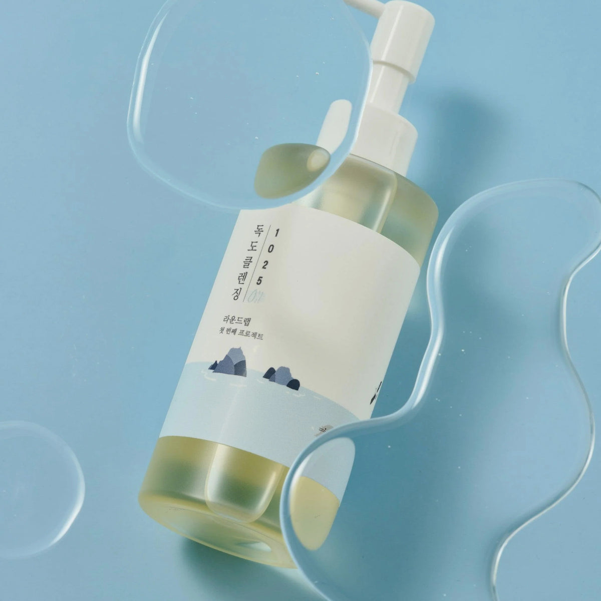 Flasche des 1025 Dokdo Cleansing Oil von Round Lab auf blauem Untergrund, umgeben von geschwungenen, transparenten Ölklecksen.