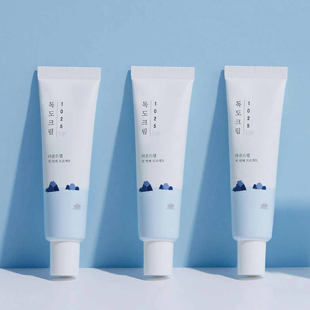 Drei Tuben der Round Lab 1025 Dokdo Eye Cream stehen aufrecht nebeneinander vor hellblauem Hintergrund.