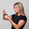 Frau in schwarzem Hemd, die ein Glas der Scandinavian Biolabs Hair Hydration Mask vor einem schlichten hellgrauen Hintergrund hält und Produkt heraus nimmt.