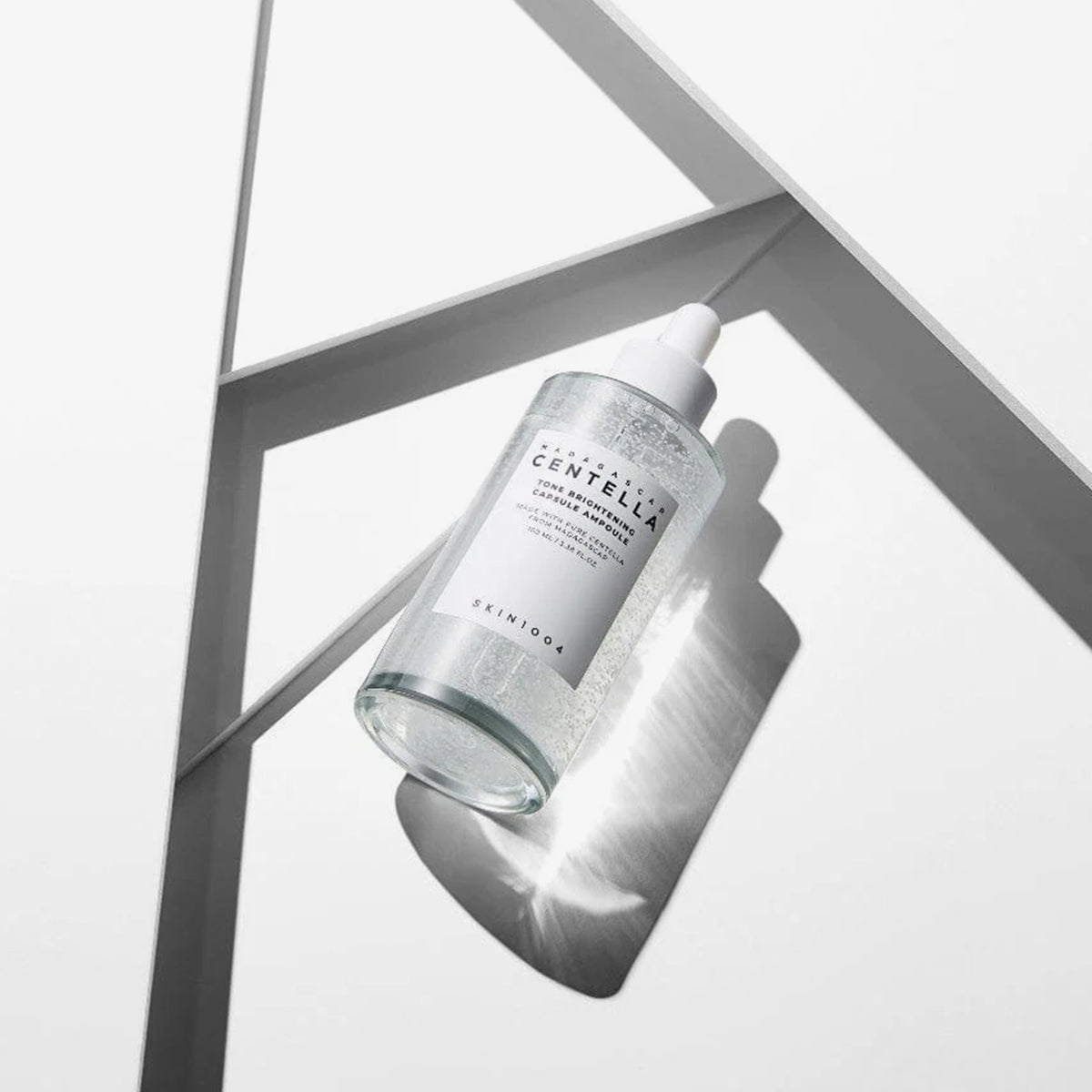 Eine durchsichtige Tropfflasche mit der Madagascar Centella Tone Brightening Capsule Ampoule wirft einen Schatten auf einen weißen geometrischen Hintergrund.