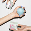 Drei Hände mit blau lackierten Nägeln halten Skinlick Cloud Cream Light in Tiegel und Verpackung – die blaue Gel-Creme läuft leicht über den Rand.