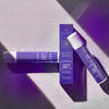Zwei violette Solar II SPF50-Flaschen und eine Schachtel, auf einer strukturierten Oberfläche unter dramatischer Beleuchtung platziert.