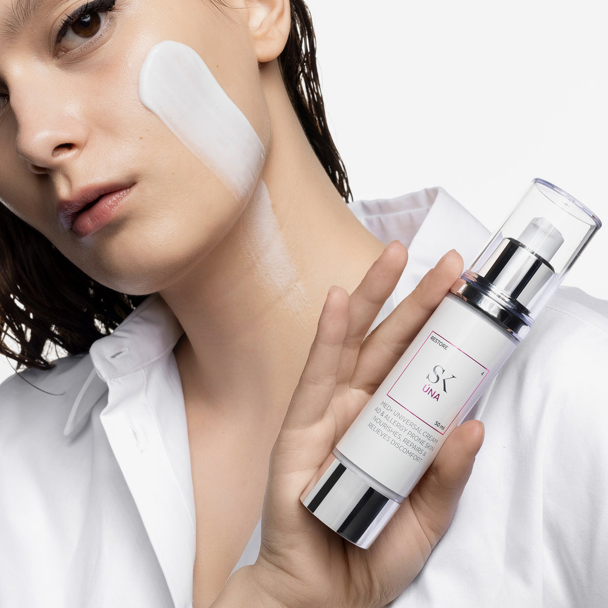 Frau mit aufgetragener Creme auf der Wange hält den Skintegra UNA Moisturizer in der Hand.