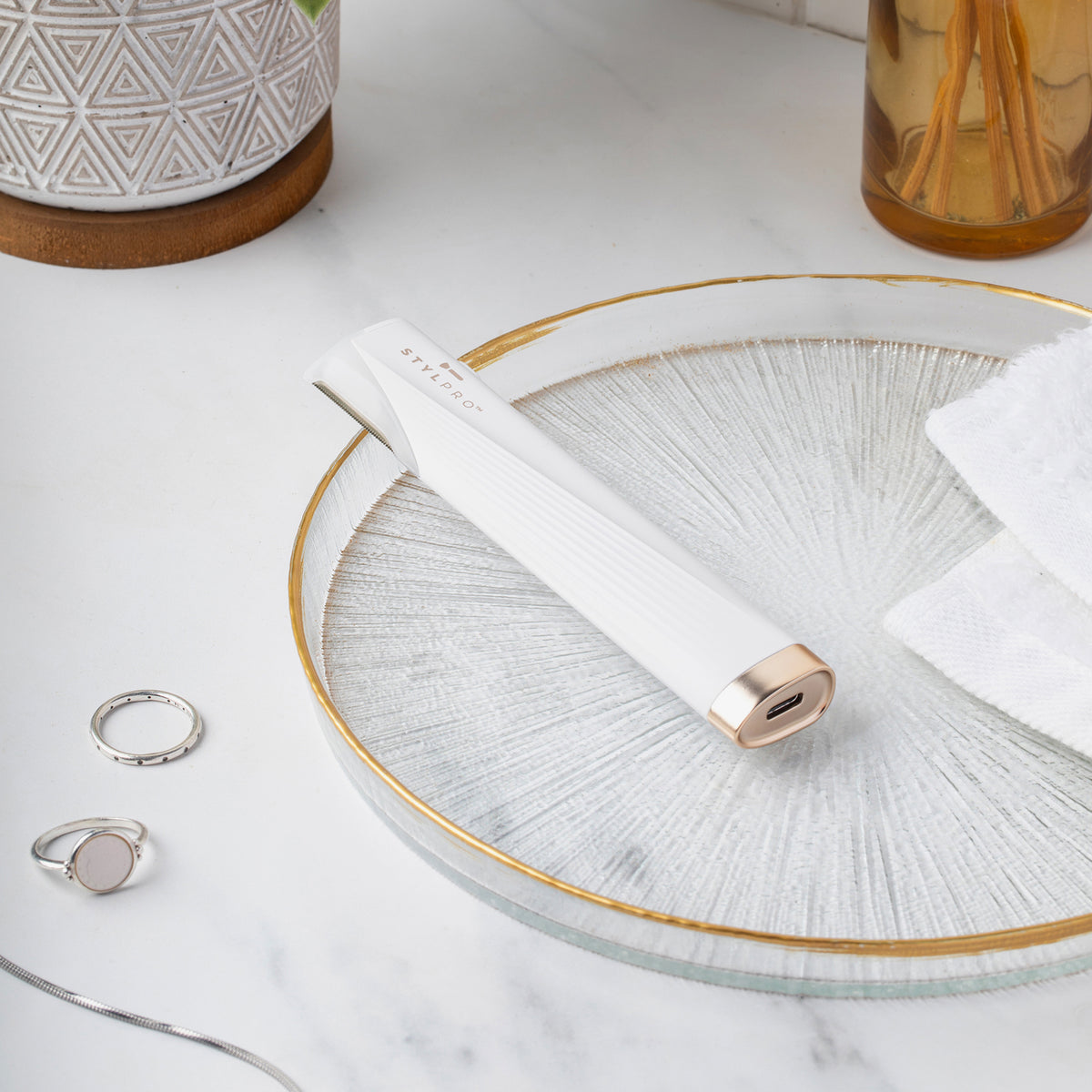 Weißes STYLPRO Dermaplaning-Gerät mit goldenen Akzenten liegt auf dekorativem Glasteller, umgeben von Schmuck und Handtuch.