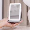Eine Hand hält den STYLPRO LED Mini Mirror, einen kompakten beleuchteten Spiegel mit klappbarem Ständer, auf einer beigen Oberfläche.
