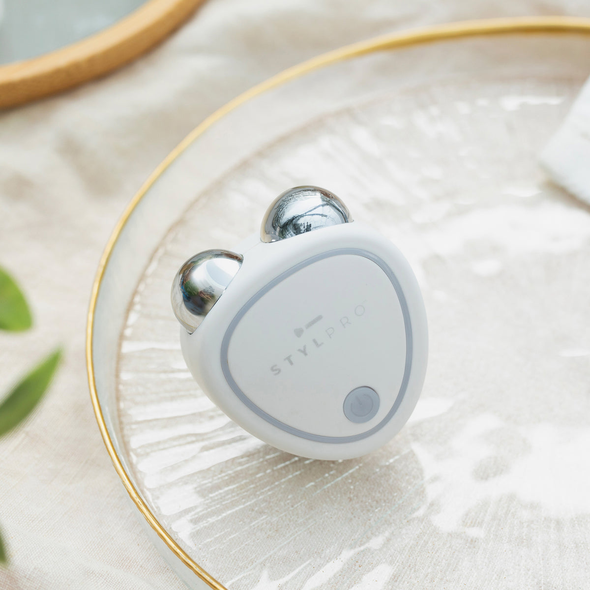Weißes STYLPRO Microcurrent-Gerät mit zwei silbernen Massagekugeln liegt auf einem Glasteller mit goldfarbenem Rand, dekorativ platziert.