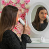 Eine Frau verwendet das Pure Red LED Light Therapy Facial Device auf ihrem Gesicht, während sie in einen Spiegel schaut; rosa und weiße Blumen im Hintergrund.
