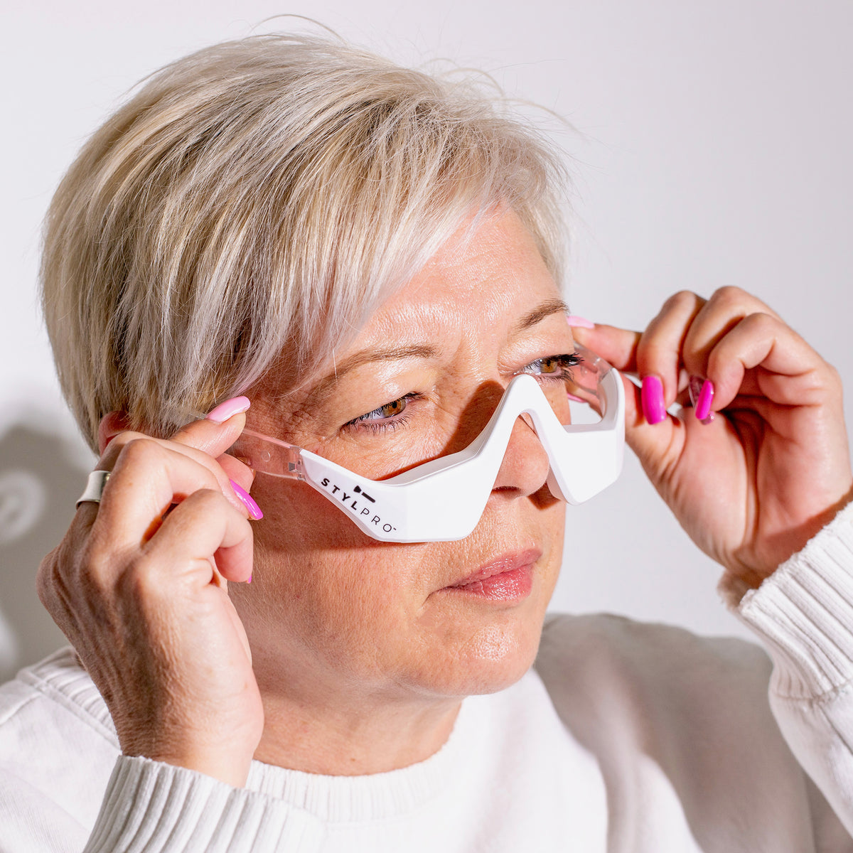 Frau mit kurzem, grauem Haar setzt weiße STYLPRO-Brille auf – ein Beauty-Tool mit Lichttherapie-Funktion für die Augenpartie.