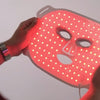 Person hält die durchleuchtete STYLPRO LED-Gesichtsmaske mit rot leuchtenden LEDs, die gleichmäßig über die Maske verteilt sind.