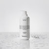 Eine Flasche AHA Smoothing Body Lotion steht mit Wassertropfen auf einer nassen Oberfläche.