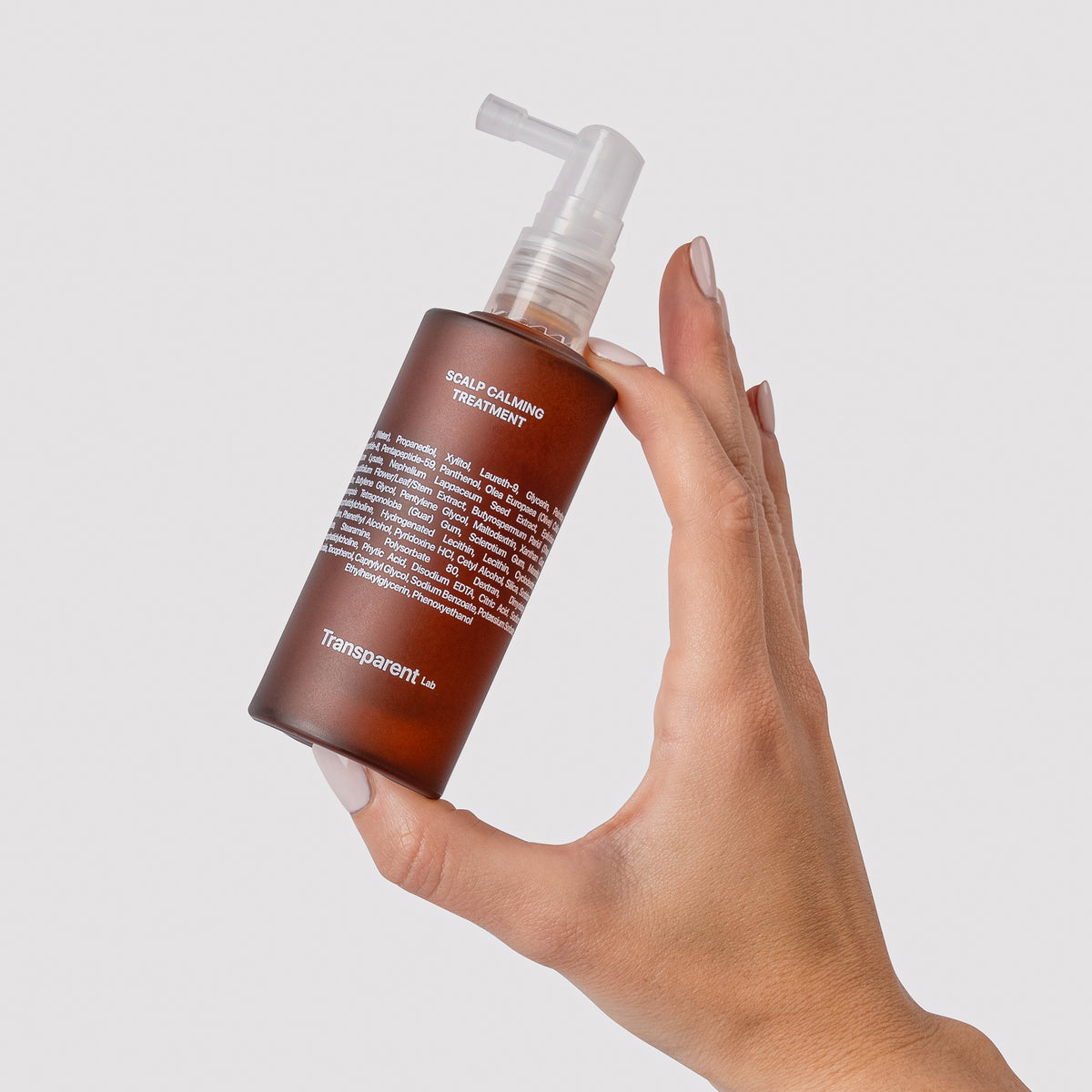 Hand hält eine braune Sprühflasche  des Scalp Calming Treatments auf weißem Hintergrund.