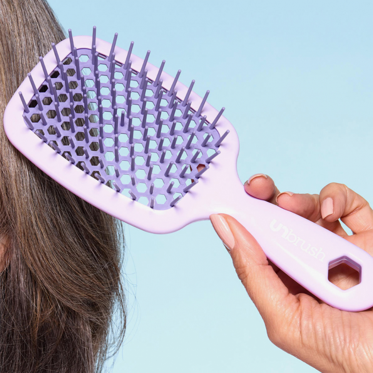 Eine Hand zeigt die lavendelfarbene Detangeling Hair Brush von UNbrush vor einem blauen Hintergrund.