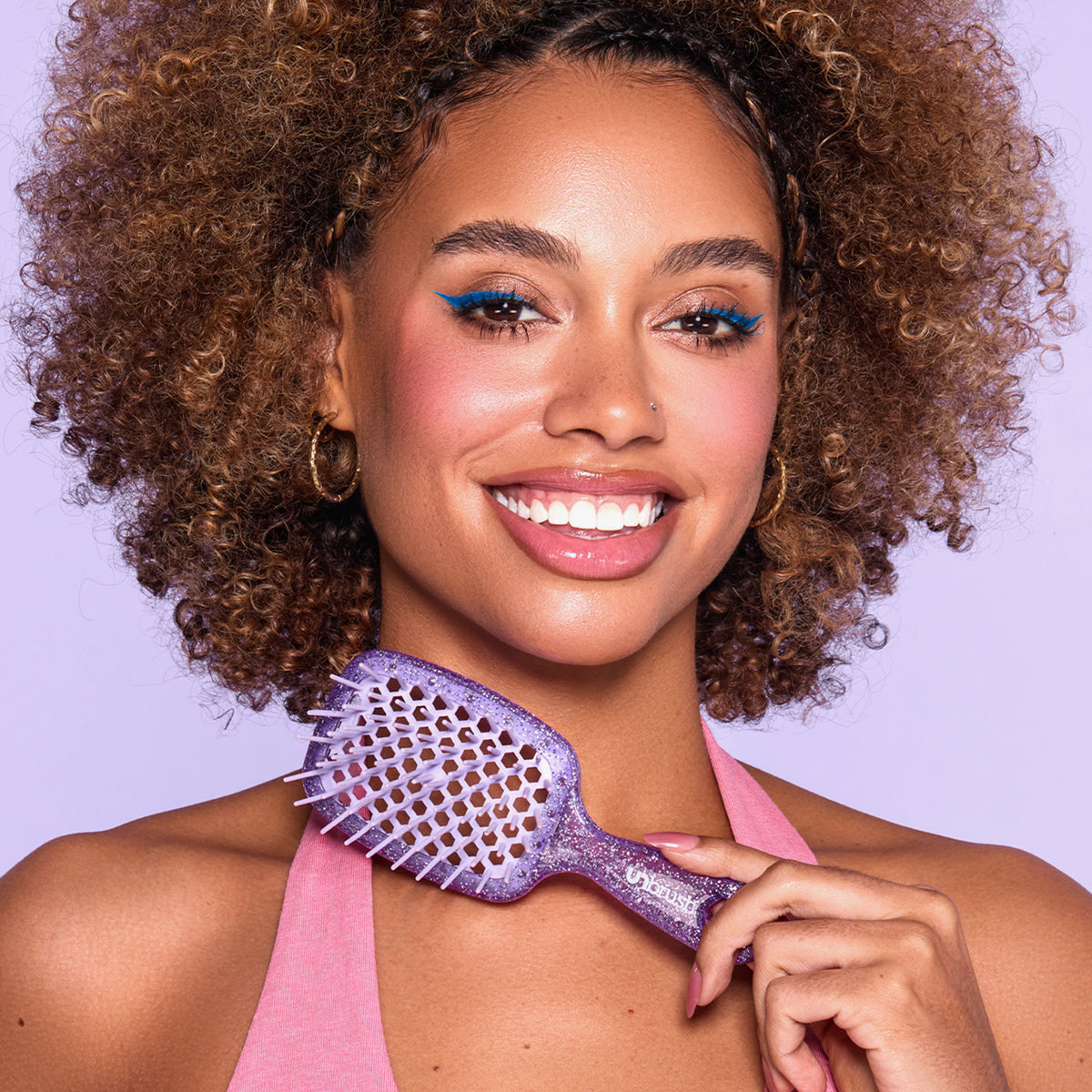 Eine lächelnde Frau mit lockigem Haar hält vor einem lila Hintergrund die lila glitzernde UNbrush Detangling Hair Brush Mini in der Hand. 